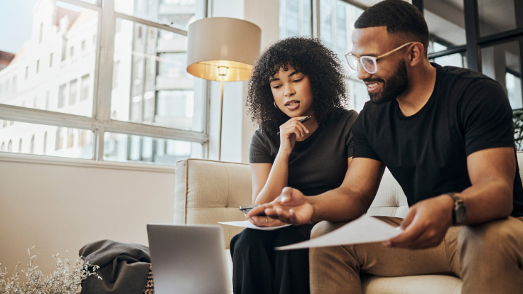 Couple sitting on sofa point at a laptop screen while shuffling papers.