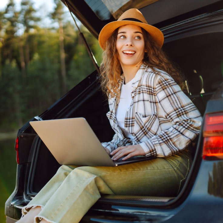 A  young woman views her laptop while siting in the back of an SUV that's parked in a wooded environment.